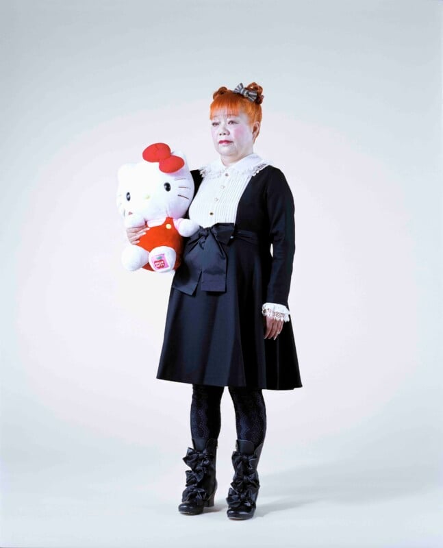 A person with bright orange hair styled in buns stands against a plain background, wearing a black dress with a white lace collar and holding a large Hello Kitty plush toy.