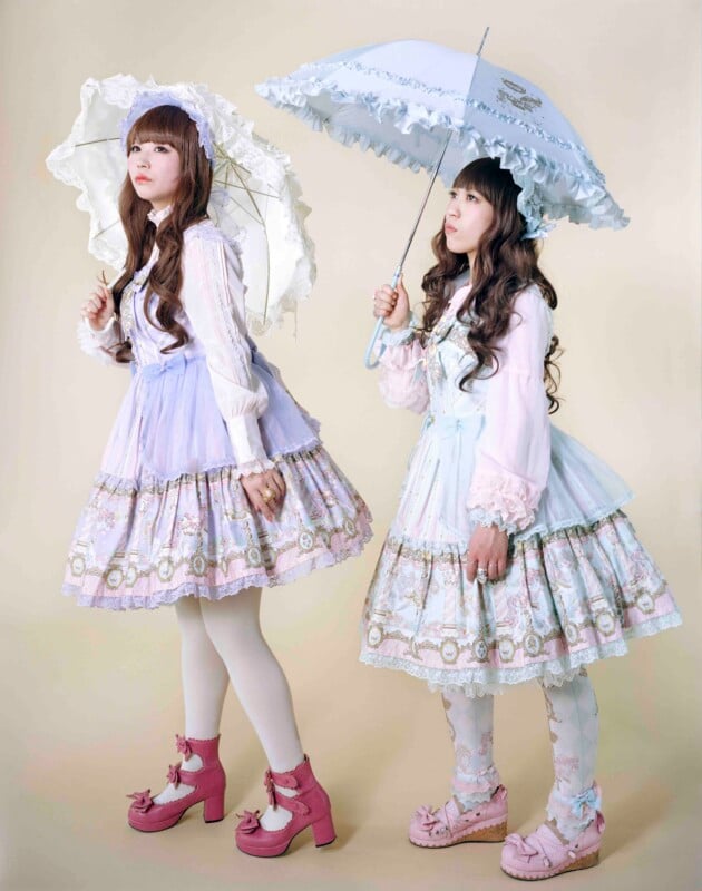 Two young women in pastel Lolita-style dresses and headwear pose against a plain background, each holding a decorative parasol.