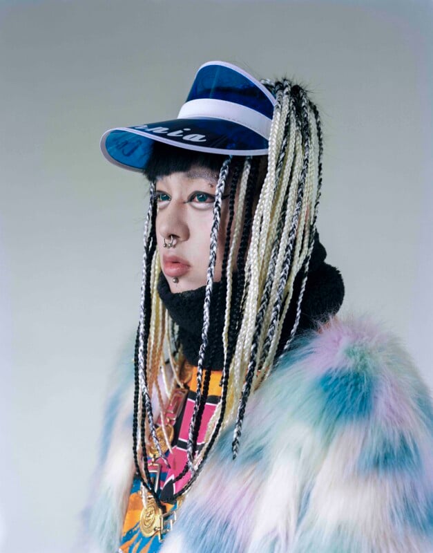 A person with long, black and white braided hair wears a blue translucent visor, a black turtleneck, and a multi-colored faux fur coat.