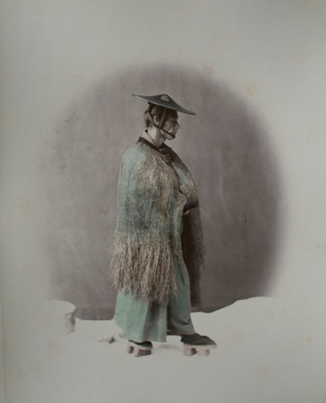A man in traditional Japanese attire, including a straw rain cape, wide-brimmed hat, and wooden sandals, stands in profile against a plain background.