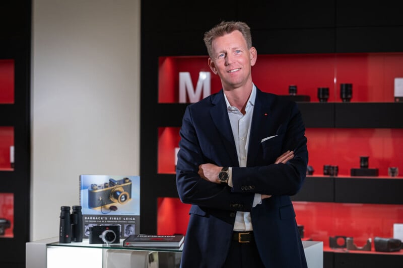 A man in a dark suit and white shirt stands with his arms crossed in front of a display shelf featuring camera equipment and a book titled Barnack's First Leica.
