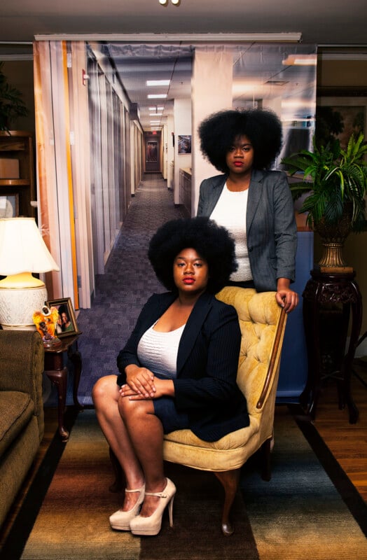 Two Black women with large afros pose for a portrait in front of a backdrop depicting an office hallway, one seated in a yellow chair and the other standing behind her.