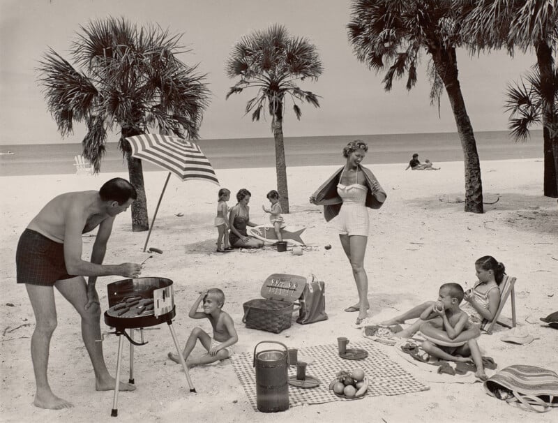 A family enjoys a beach picnic with a man grilling hot dogs, a woman draping a towel over her shoulders, and children playing or eating on a checkered blanket.