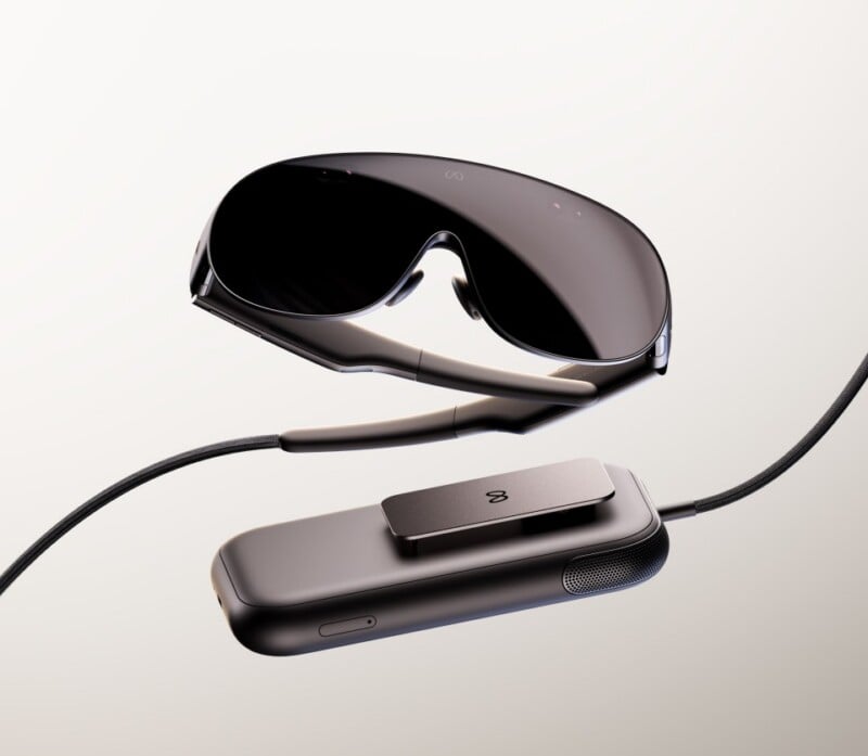 A pair of sleek, dark-tinted smart glasses connected by a cable to a rectangular, handheld control unit with a clip.