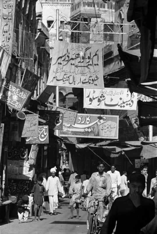 A crowded, narrow street in a South Asian city features numerous hanging signs with Urdu text, pedestrians, and a person riding a bicycle.