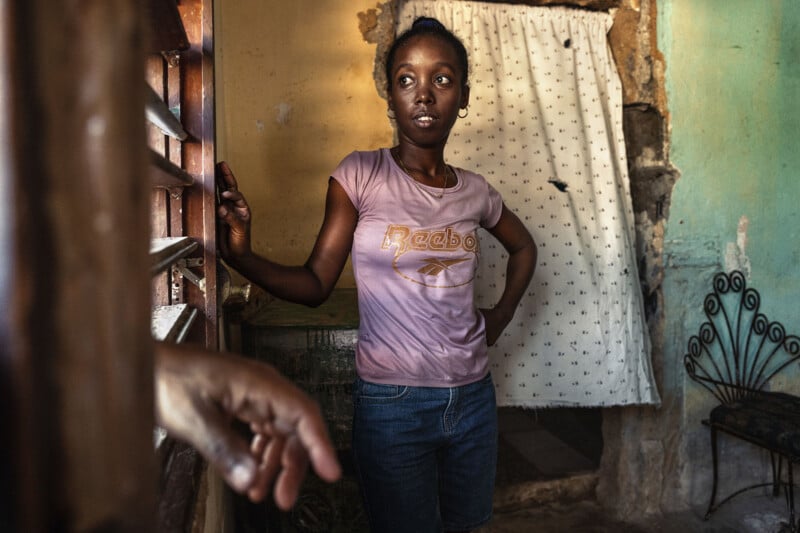 A young woman in a pink Reebok t-shirt and blue jeans stands indoors, looking toward a window while a hand reaches into the frame.