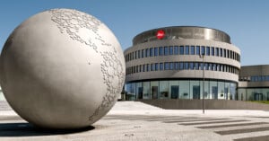 A large, spherical sculpture with a textured map surface stands on a paved plaza in front of a modern, circular building with the Leica logo.