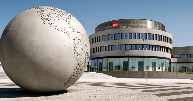 A large, spherical sculpture with a textured map surface stands on a paved plaza in front of a modern, circular building with the Leica logo.
