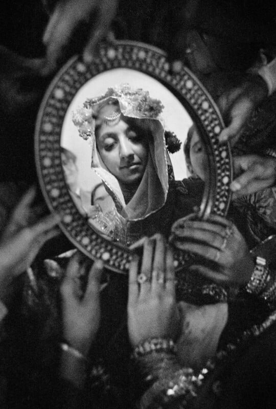 A black-and-white close-up shows a person wearing a traditional head covering, their face reflected in an ornate, circular hand mirror held by multiple hands.