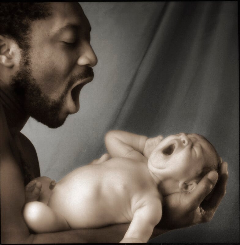 A man with a beard holds a newborn baby in his hands, and both are yawning with their mouths wide open.