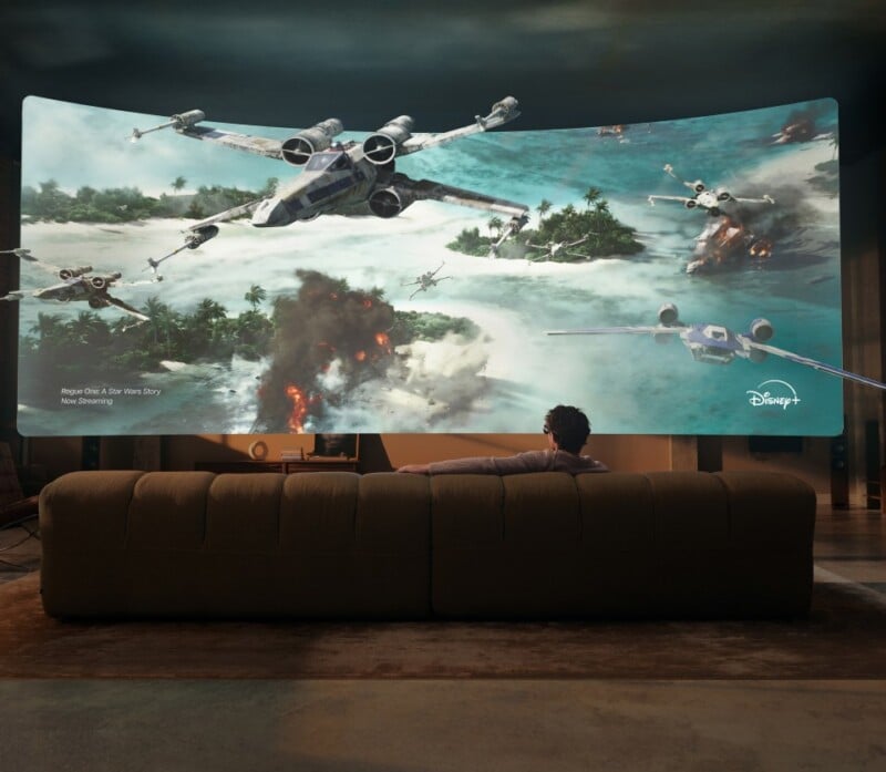 A person sits on a sofa watching a large, curved screen displaying a Star Wars space battle scene with X-wing starfighters.