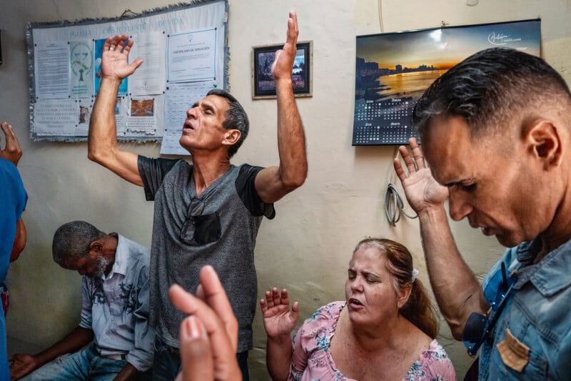 A group of people pray indoors with their hands raised, while a man sits nearby with his head bowed.