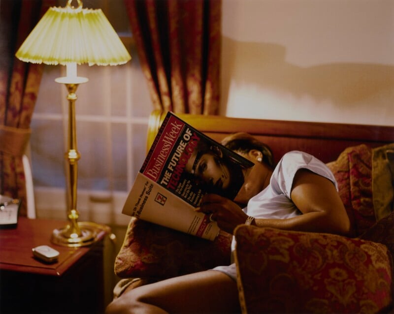 A person reclines on a patterned sofa, holding a BusinessWeek magazine in front of their face, next to a lit table lamp.