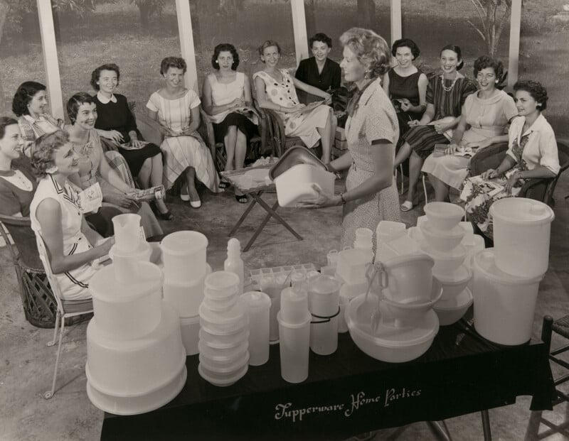 A woman demonstrates Tupperware products to a group of women seated in a semi-circle at a home party.