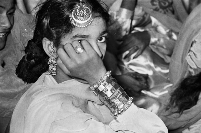 A young person with dark hair and traditional jewelry, including a maang tikka and bangles, covers part of their face with their hand.