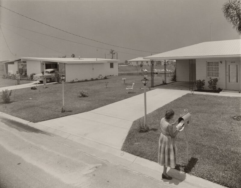 A woman in a dress checks her mailbox in a suburban neighborhood while a man mows the lawn of a nearby house.