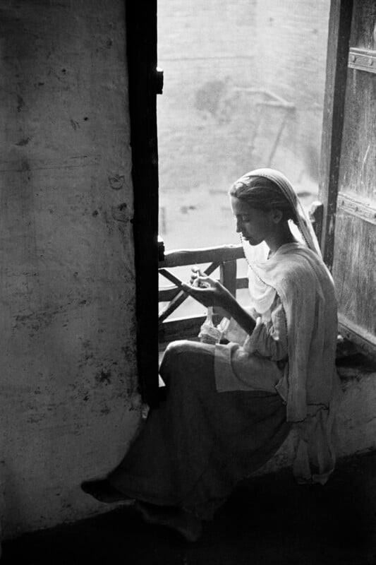 A woman in a headscarf sits by a window, focused on sewing or needlework in her hands.