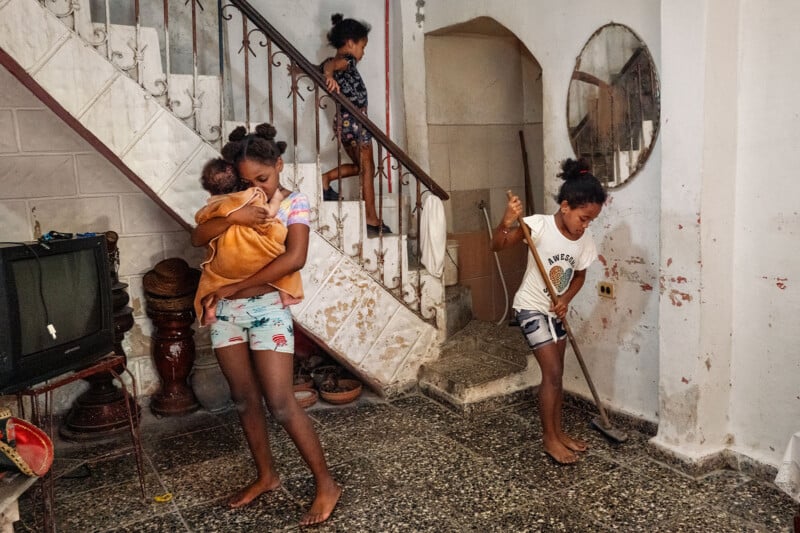 Three children are in a room with a staircase; one girl holds a baby, another sweeps the floor, and a third walks down the stairs.