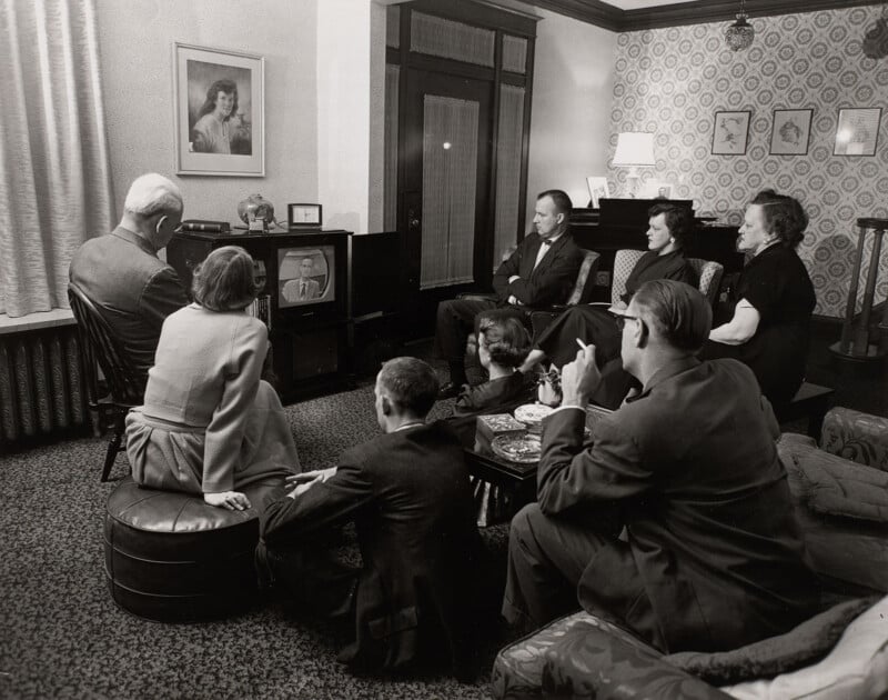 A group of seven people sit in a living room watching a man on a television set.