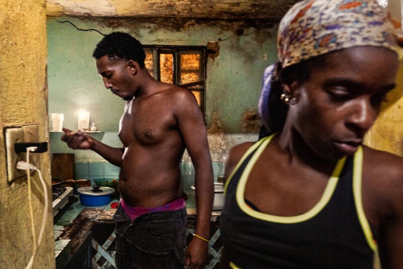 A shirtless man stands in a kitchen while a woman wearing a headscarf and a black tank top stands in the foreground.