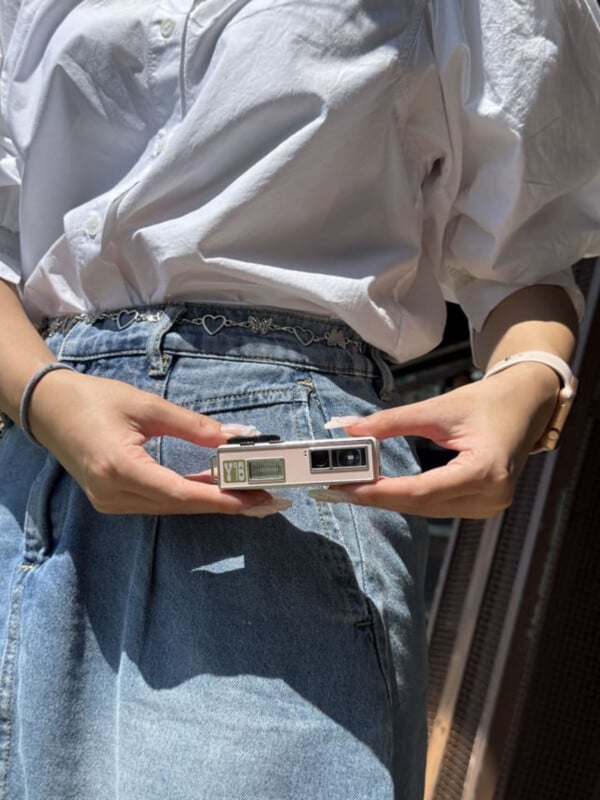 A person wearing a white button-down shirt and denim skirt holds a small, rectangular, light pink camera with a "Y16" label in their hands.