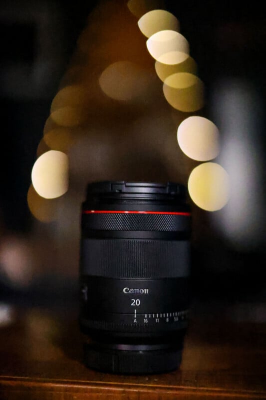 A black Canon camera lens with a red ring stands upright on a wooden surface against a background of blurred, golden bokeh lights.