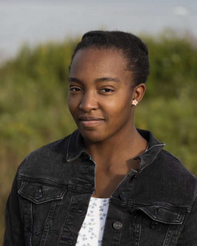 A young woman with short, dark hair and a light floral top under a black denim jacket looks toward the camera against a blurred green background.