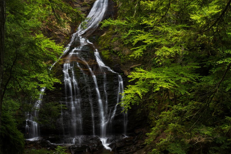 A tall, tiered waterfall cascades down a dark, mossy rock face surrounded by lush green forest foliage.