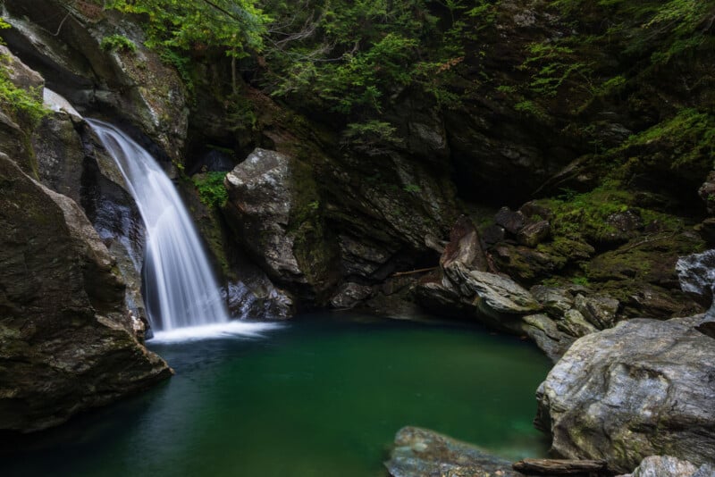 A waterfall cascades into a deep, green pool surrounded by mossy rock formations and dense forest foliage.