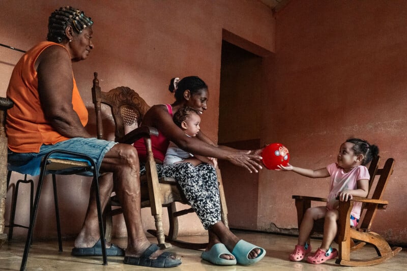 An older woman, a younger woman holding a baby, and a young girl sitting in a rocking chair pass a red ball to each other.