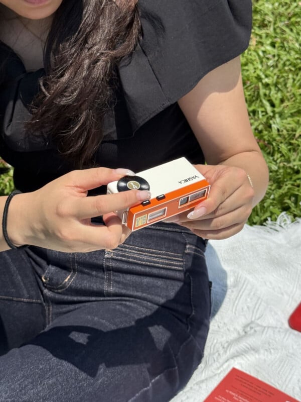 A person with manicured nails holds a small, rectangular white and orange Yashica Y16 camera while sitting on a blanket outdoors.