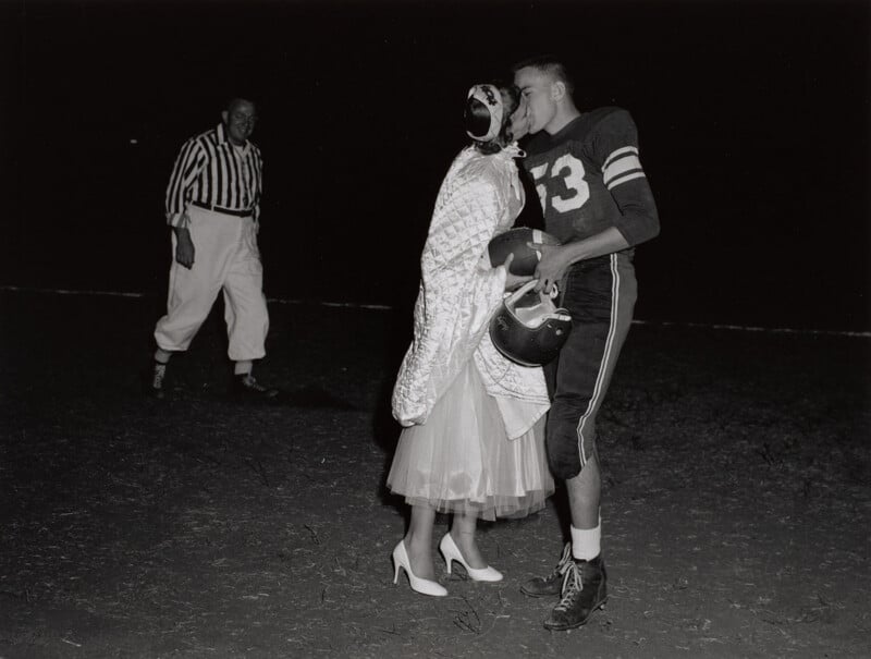 A football player in uniform kisses a woman in a formal dress and heels on a field at night, while a referee in a striped shirt watches in the background.