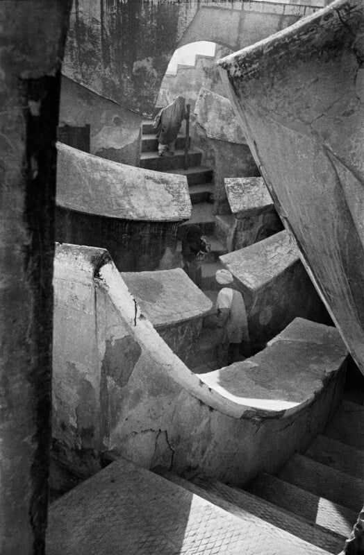A high-angle, black-and-white shot shows people walking up stone stairs within the geometric, weathered structures of an observatory.