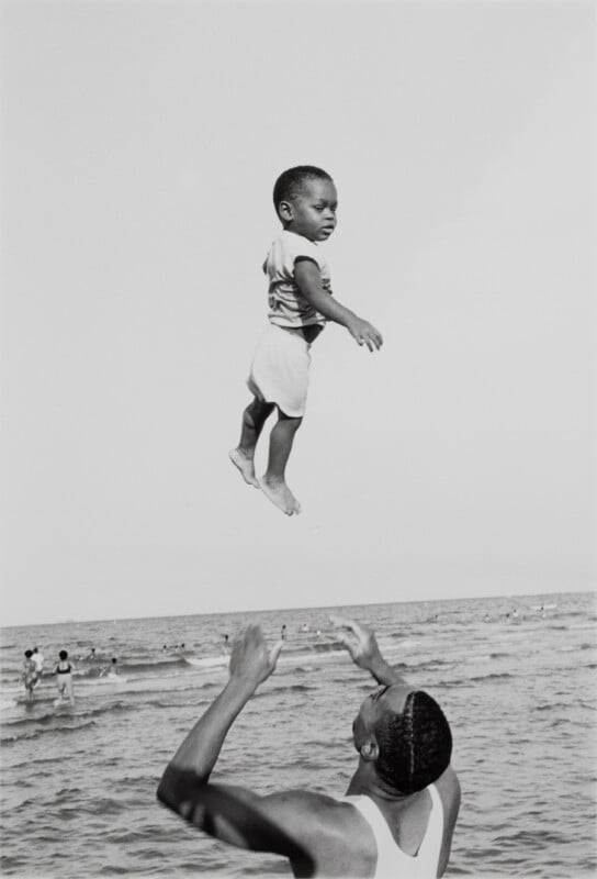 A man in a white tank top tosses a young child into the air above the ocean, with other people swimming in the background.