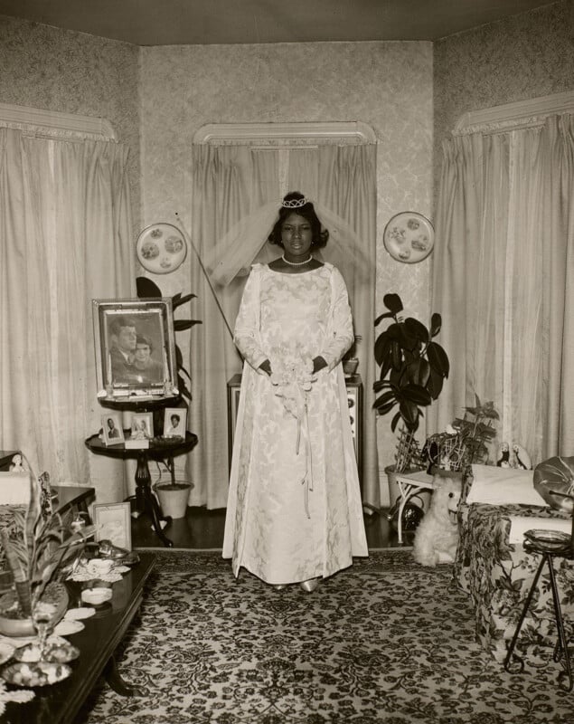 A Black bride in a long-sleeved white wedding dress and veil stands in a living room, with a framed portrait of John and Jackie Kennedy on a side table nearby.