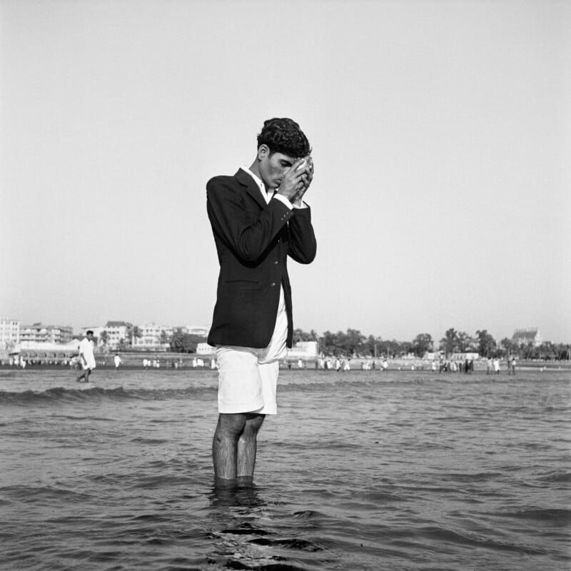 A man in a dark blazer and white shorts stands in shallow ocean water, holding a seashell to his face with both hands.