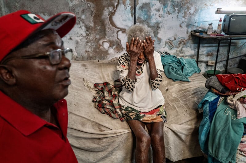 A man in a red cap and shirt stands in the foreground while an elderly woman sits on a bed behind him with her hands covering her face.
