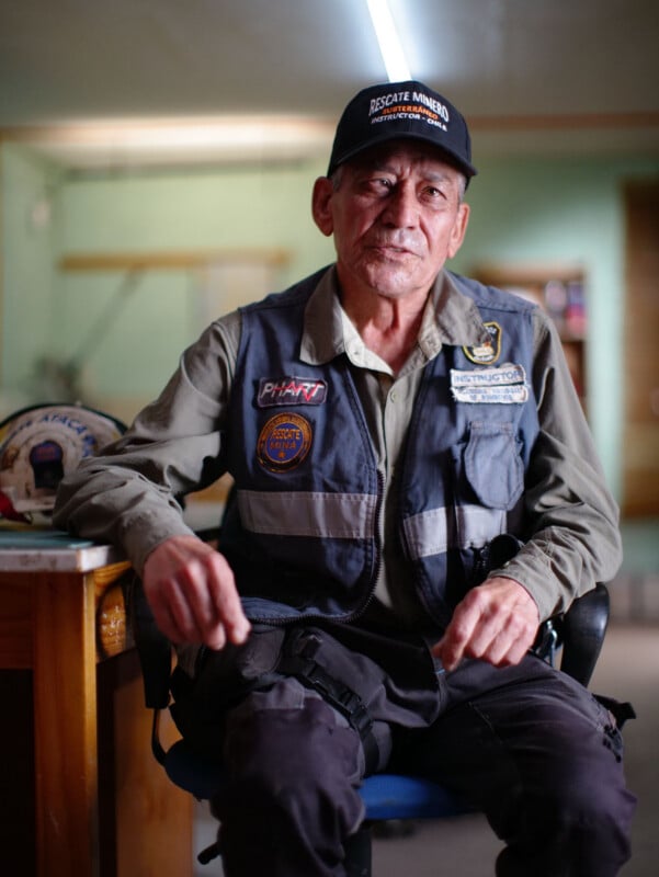 An older man wearing a dark cap and a vest with various patches sits in a chair, looking directly at the camera.