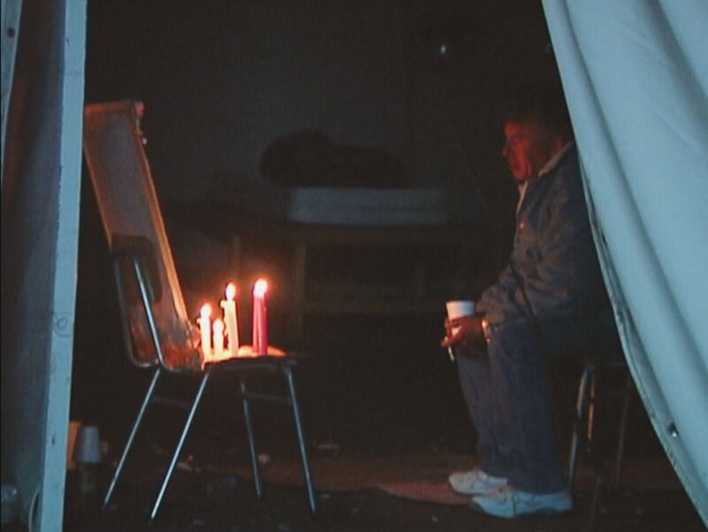 A man sits on a chair in a dimly lit space, holding a cup and facing a chair holding several lit candles.