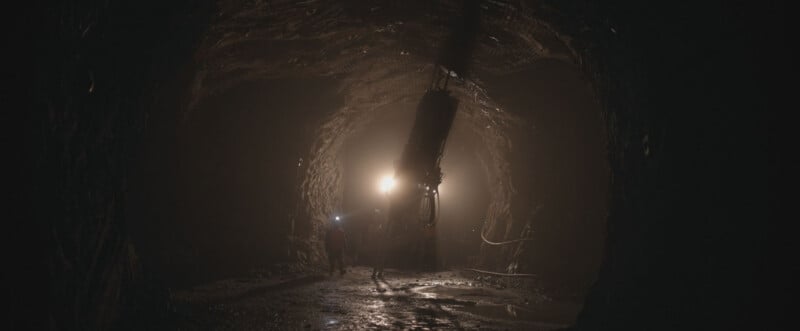 A person wearing a headlamp walks through a dark, rocky mine tunnel toward a large, illuminated drilling machine.