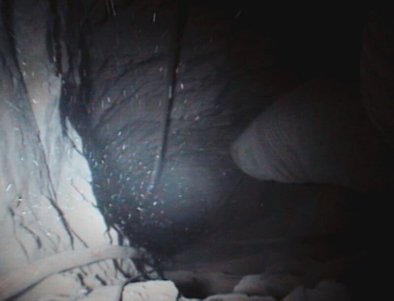 A dark, cavernous space with rough, textured rock walls illuminated by a light source, with a blurry object protruding from the right side.