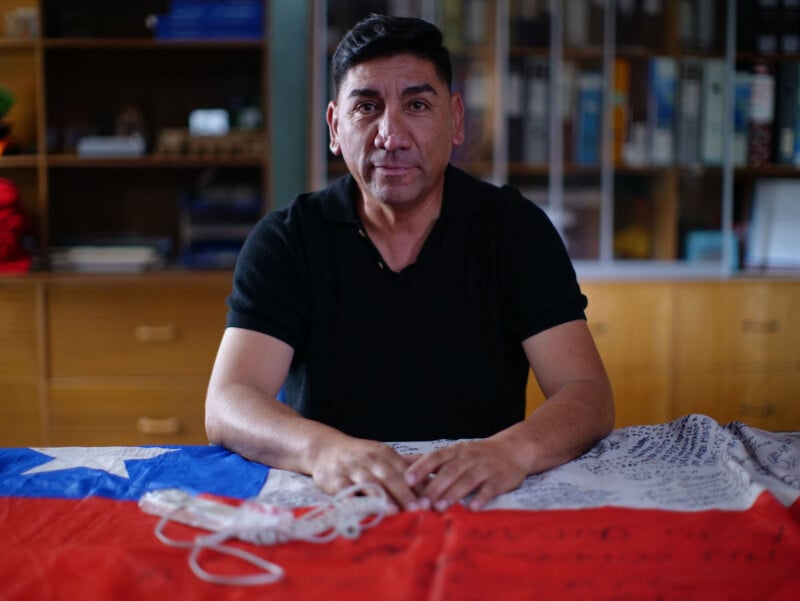 A middle-aged man with dark hair sits at a table draped with a Chilean flag covered in handwritten signatures.