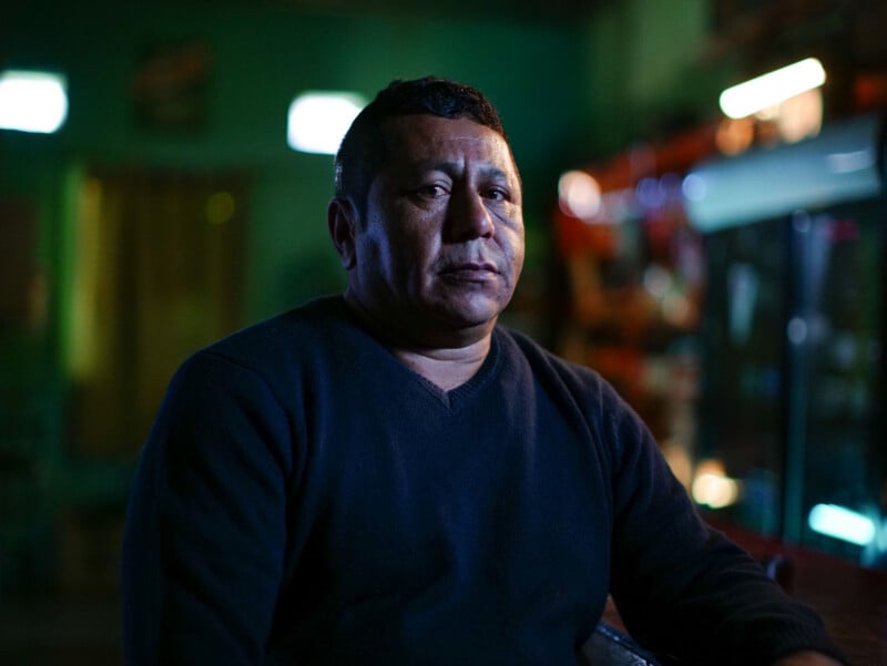 A middle-aged man with short dark hair and a dark V-neck sweater sits in a dimly lit room with green walls.