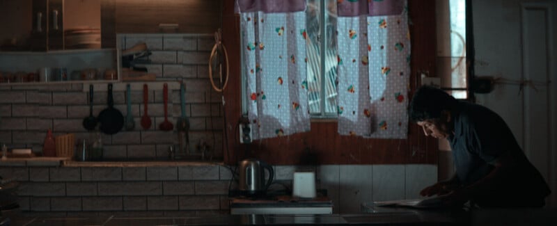 An older man leans over a kitchen counter, looking down at a document, in a dimly lit kitchen with patterned curtains and hanging utensils.