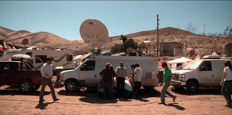 A group of people gathers around a white satellite news van in a desert landscape with mountains in the background.