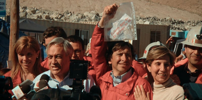 A group of people, including former Chilean President Sebastián Piñera, stand before microphones while a man in a red jacket holds up a plastic bag containing a note that reads "Estamos bien en el refugio los 33.