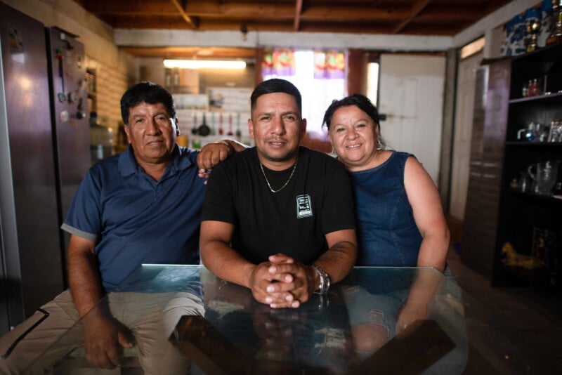 Three people sit together at a glass table in a home kitchen, smiling at the camera.