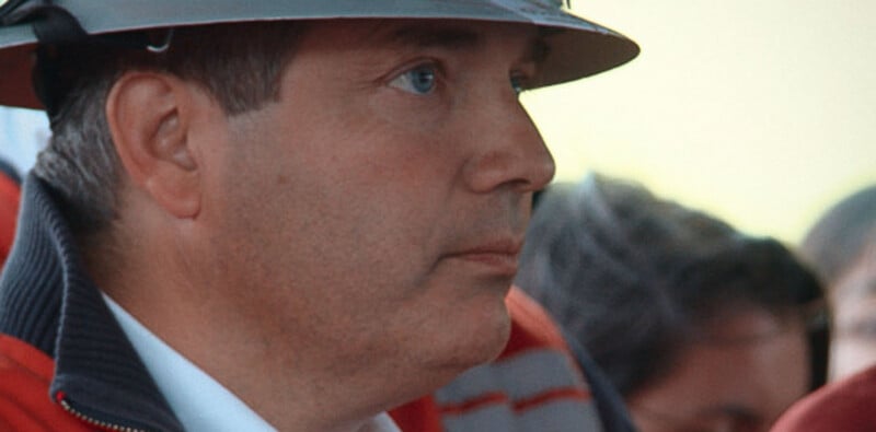A side profile of a man wearing a hard hat and a red jacket with a white collared shirt underneath.