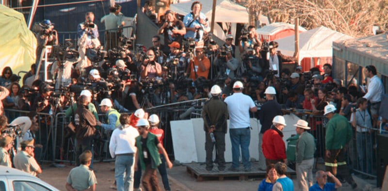 A large group of photographers and journalists with cameras and equipment stand behind metal barricades at an outdoor event.
