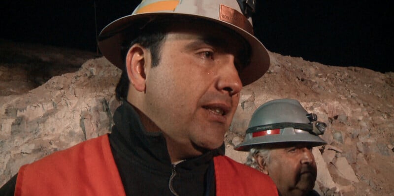 A man in a hard hat and orange safety vest speaks in front of a rocky background, with another man in a grey hard hat partially visible behind him.
